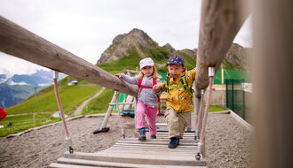 Zwei Kinder schaukeln auf einer Hängebrücke im Kleinwalsertal, umgeben von Bergen. | © Kleinwalsertal Tourismus eGen • Fotograf: Oliver Farys