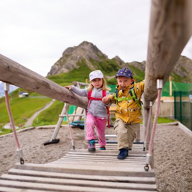 Zwei Kinder schaukeln auf einer Hängebrücke im Kleinwalsertal, umgeben von Bergen. | © Kleinwalsertal Tourismus eGen • Fotograf: Oliver Farys