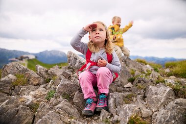 Zwei Kinder beim Wandern in den Bergen, eine junge Mädchen sitzt auf Steinen und schaut nach oben, ein älterer Junge im Hintergrund hebt die Hand zum Gruß. | © Kleinwalsertal Tourismus eGen • Fotograf: Oliver Farys