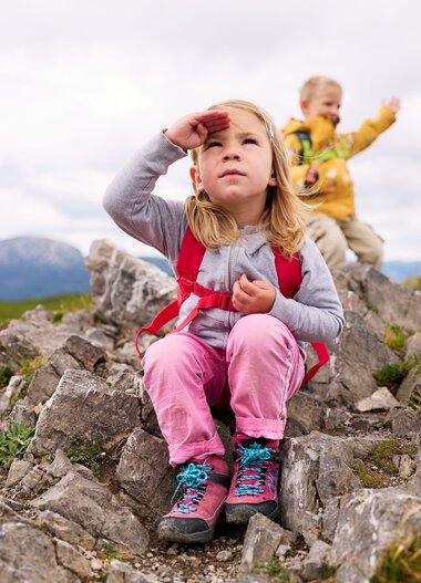 Zwei Kinder beim Wandern in den Bergen, eine junge Mädchen sitzt auf Steinen und schaut nach oben, ein älterer Junge im Hintergrund hebt die Hand zum Gruß. | © Kleinwalsertal Tourismus eGen • Fotograf: Oliver Farys