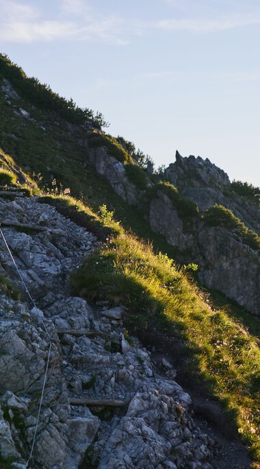 Bergpfad im Kleinwalsertal bei Sonnenaufgang, mit Steigungen und grüner Vegetation entlang der Felsen.“},{ | © Kleinwalsertal Tourismus eGen • Fotograf: Oliver Farys