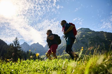 Zwei Wanderer mit Rucksack gehen in einer Berglandschaft im Sommer, gegen den Himmel fotografiert. | © Kleinwalsertal Tourismus eGen • Fotograf: Oliver Farys