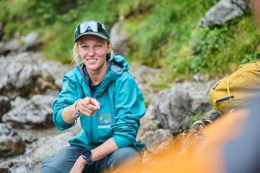 Freundliche Frau in Outdoor-Bekleidung, sitzend an einem Fluss mit Steinen und Natur im Hintergrund. | © Kleinwalsertal Tourismus eGen • Fotograf: Oliver Farys