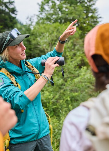 Nature guide explaining to a group during an outdoor excursion in Kleinwalsertal. The background features trees and mountains. | © Kleinwalsertal Tourismus eGen • Photographer: Oliver Farys