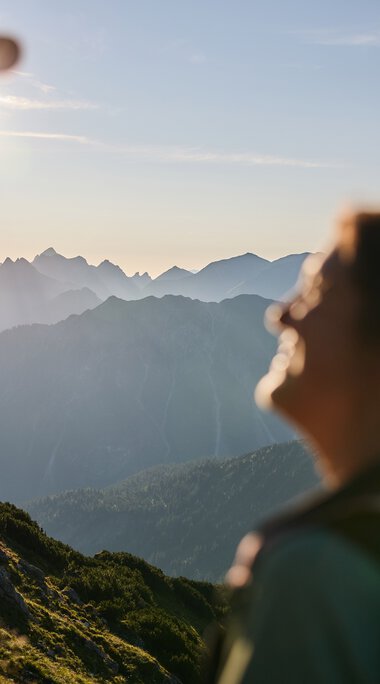 Two hikers in the mountains at sunrise in Kleinwalsertal, with mountain ranges in the background and sunlight behind one person. | © Kleinwalsertal Tourismus eGen • Photographer: Oliver Farys