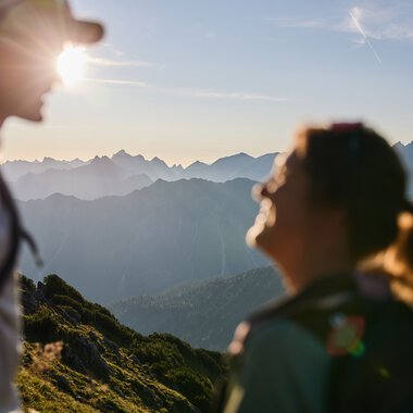Two hikers in the mountains at sunrise in Kleinwalsertal, with mountain ranges in the background and sunlight behind one person. | © Kleinwalsertal Tourismus eGen • Photographer: Oliver Farys