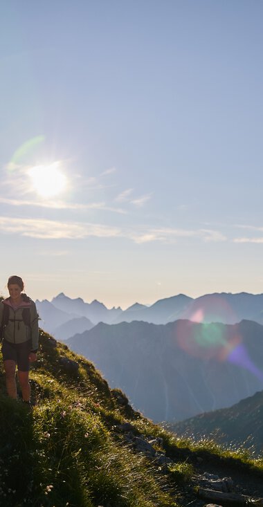 Drei Wanderer auf einem schmalen Bergpfad im Kleinwalsertal bei Sonnenaufgang mit Berggipfeln im Hintergrund. | © Kleinwalsertal Tourismus eGen • Fotograf: Oliver Farys