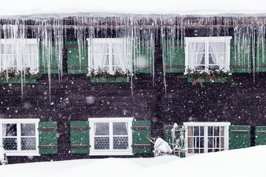 Schneebedecktes Haus mit grünen Fensterläden und Eiszapfen im Winter bei starkem Schneefall. | © Kleinwalsertal Tourismus eGen • Fotograf: Oliver Farys