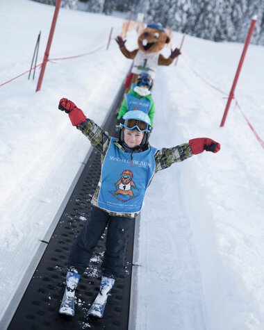 Ein Kind auf Skiern fährt stolz auf einer Schneise, begleitet von einem großen, lachenden Maskottchen im Hintergrund im Winterskiurlaub. | © Kleinwalsertal Tourismus eGen • Fotograf: Oliver Farys