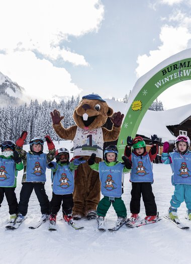 Kinderskikurs mit Kindern und einer Skimaske im Schnee, unter einem Bogen mit dem Schriftzug 'Burmi Winterwelt' in einer verschneiten Bergregion. | © Kleinwalsertal Tourismus eGen • Fotograf: Oliver Farys