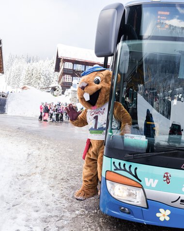 Maskottchen im Winter vor einem Bus, Schnee, Kinder im Hintergrund, Skifahrzeug, Winterkleidung. | © Kleinwalsertal Tourismus eGen • Fotograf: Oliver Farys