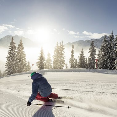 Ein Kind beim Skifahren auf einer frisch präparierten Piste in den verschneiten Bergen, umgeben von Tannen und bei sonnigem Himmel. | © Kleinwalsertal Tourismus eGen • Fotograf: Oliver Farys