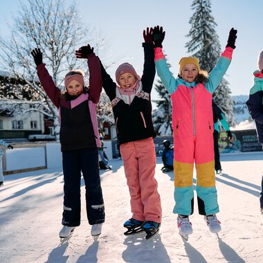 Kinder beim Eislaufen auf einem zugefrorenen See im Winter, fröhlich mit erhobenen Armen, umgeben von verschneiten Bäumen und anderen Eisläufern. | © Kleinwalsertal Tourismus eGen • Fotograf: Oliver Farys
