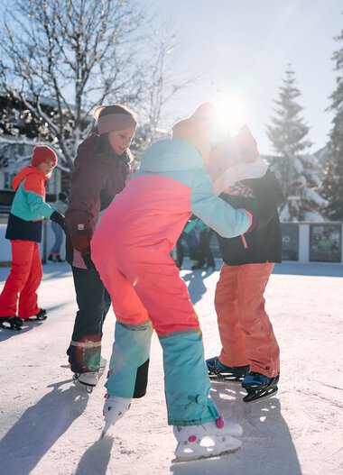 Kinder beim Eislaufen auf einem Eisplatz im Winter, umgeben von Schneefeldern und Bäumen, mit einem Gebäude im Hintergrund. | © Kleinwalsertal Tourismus eGen • Fotograf: Oliver Farys