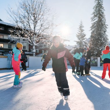 Kinder beim Eislaufen auf einem Eisparkplatz im Winter, umgeben von Bäumen und Gebäuden, sonniger Tag. | © Kleinwalsertal Tourismus eGen • Fotograf: Oliver Farys