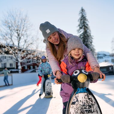 Kinder beim Eislaufen auf einem winterlichen Eisplatz in Riezlern im Kleinwalsertal, Sonnenlicht und schneebedeckte Bäume im Hintergrund. | © Kleinwalsertal Tourismus eGen • Fotograf: Oliver Farys