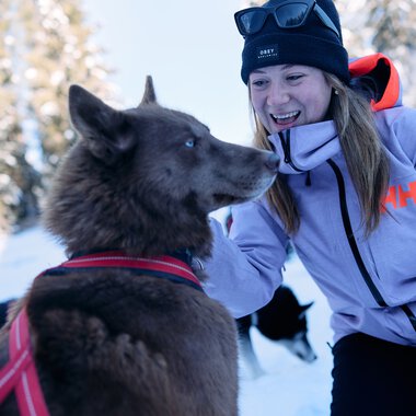 Frau im Winter-Outfit spielt mit einem Husky in verschneiter Winterlandschaft. | ©  Kleinwalsertal Tourismus eGen • Fotograf: Oliver Farys