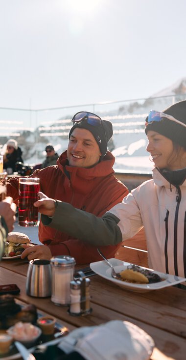 Drei Freunde beim Mittagessen im Freien mit Blick auf schneebedeckte Berge im Kleinwalsertal, sie stoßen mit Getränken an. | © Kleinwalsertal Tourismus eGen • Fotograf: Oliver Farys