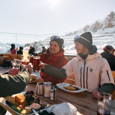 Drei Freunde beim Mittagessen im Freien mit Blick auf schneebedeckte Berge im Kleinwalsertal, sie stoßen mit Getränken an. | © Kleinwalsertal Tourismus eGen • Fotograf: Oliver Farys