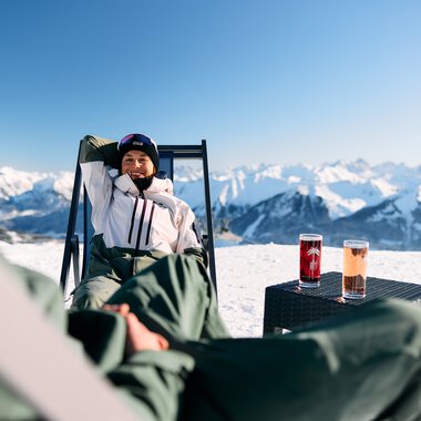Person entspannt auf einer Sonnenliege im Schnee mit Blick auf die verschneiten Berge, Getränke auf einem Tisch neben sich. | © Kleinwalsertal Tourismus eGen • Fotograf: Oliver Farys