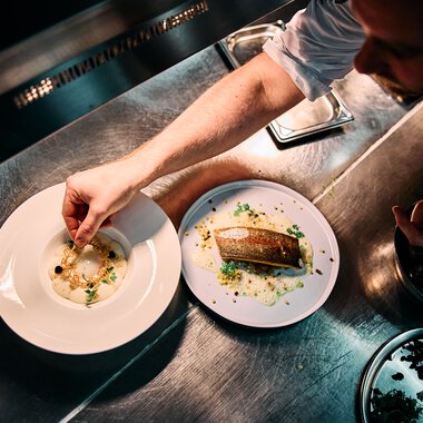 A chef garnishes a dish in a professional kitchen, on a metallic work surface, with two white plates and modern kitchen tools. | © Kleinwalsertal Tourismus eGen • Photographer: Oliver Farys