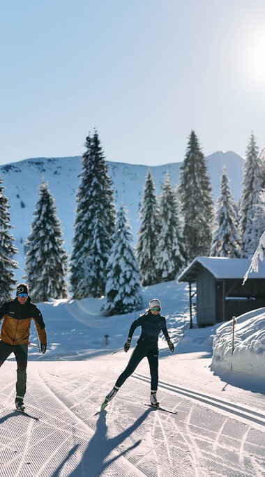 Zwei Skifahrer ziehen ihre Spuren im verschneiten Winterwald beim Langlaufen im Kleinwalsertal, Sonnenschein und klare Sicht. | ©  Kleinwalsertal Tourismus eGen • Fotograf: Oliver Farys