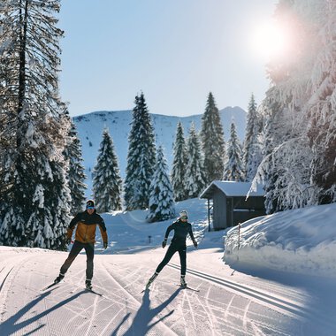 Two cross-country skiers enjoying skiing in a snowy winter forest in Kleinwalsertal under sunny skies. | © Kleinwalsertal Tourismus eGen • Photographer: Oliver Farys