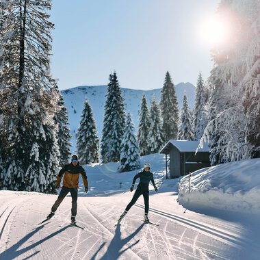 Two cross-country skiers enjoying skiing in a snowy winter forest in Kleinwalsertal under sunny skies. | © Kleinwalsertal Tourismus eGen • Photographer: Oliver Farys