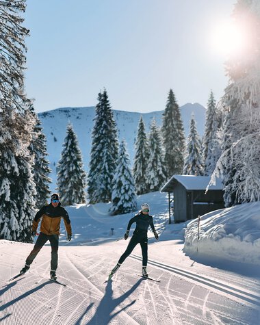 Zwei Skifahrer ziehen ihre Spuren im verschneiten Winterwald beim Langlaufen im Kleinwalsertal, Sonnenschein und klare Sicht. | ©  Kleinwalsertal Tourismus eGen • Fotograf: Oliver Farys