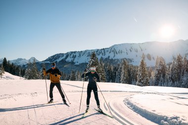 Zwei Skilangläufer in winterlicher Landschaft im Kleinwalsertal, umgeben von schneebedeckten Bäumen und Bergen, bei Sonnenschein. | ©  Kleinwalsertal Tourismus eGen • Fotograf: Oliver Farys