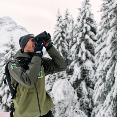 Man in winter clothing with binoculars in snowy forest in the Alps, likely a ranger or nature guide. | © Kleinwalsertal Tourismus eGen • Photographer: Oliver Farys