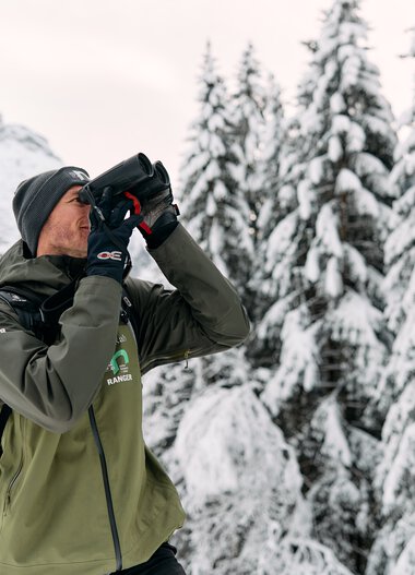 Mann mit Winterbekleidung und Fernglas in verschneitem Waldgebiet in den Alpen, wahrscheinlich ein Ranger oder Naturführer. | © Kleinwalsertal Tourismus eGen • Fotograf: Oliver Farys