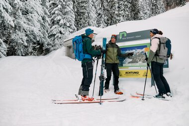 Drei Skitourengeher vor einem Informationsschild im verschneiten Wald bei Kleinwalsertal, Winter 2024. | © Kleinwalsertal Tourismus eGen • Fotograf: Oliver Farys