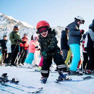 Kleiner Junge beim Skifahren mit einem lachenden Gesicht, umgeben von anderen Skifahrern in einer Winterlandschaft mit Bergen. | © Kleinwalsertal Tourismus eGen • Fotograf: Oliver Farys