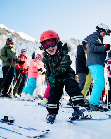 Kleiner Junge beim Skifahren mit einem lachenden Gesicht, umgeben von anderen Skifahrern in einer Winterlandschaft mit Bergen. | © Kleinwalsertal Tourismus eGen • Fotograf: Oliver Farys