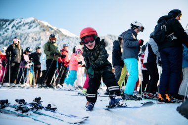 Kleiner Junge beim Skifahren mit einem lachenden Gesicht, umgeben von anderen Skifahrern in einer Winterlandschaft mit Bergen. | © Kleinwalsertal Tourismus eGen • Fotograf: Oliver Farys