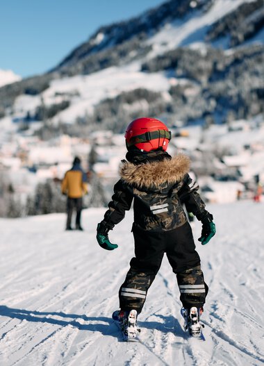 Ein Kind beim Skifahren auf einer Piste im Schnee, umgeben von Bergen und anderen Skifahrern, in winterlicher Landschaft. | © Kleinwalsertal Tourismus eGen • Fotograf: Oliver Farys