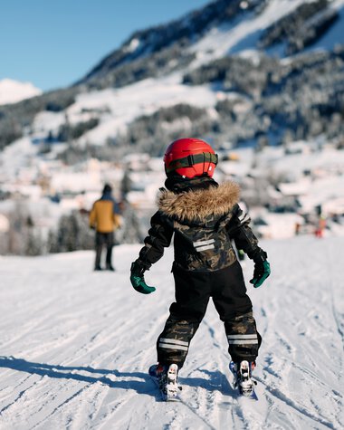 Ein Kind beim Skifahren auf einer Piste im Schnee, umgeben von Bergen und anderen Skifahrern, in winterlicher Landschaft. | © Kleinwalsertal Tourismus eGen • Fotograf: Oliver Farys
