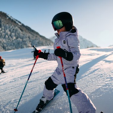 Kleines Kind beim Skifahren im Schneeland, Sonnenlicht, Berge im Hintergrund, trägt Helm und Skiausrüstung. | © Kleinwalsertal Tourismus eGen • Fotograf: Oliver Farys