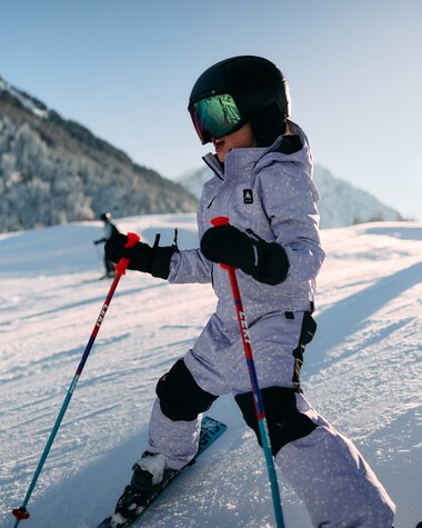 Kleines Kind beim Skifahren im Schneeland, Sonnenlicht, Berge im Hintergrund, trägt Helm und Skiausrüstung. | © Kleinwalsertal Tourismus eGen • Fotograf: Oliver Farys