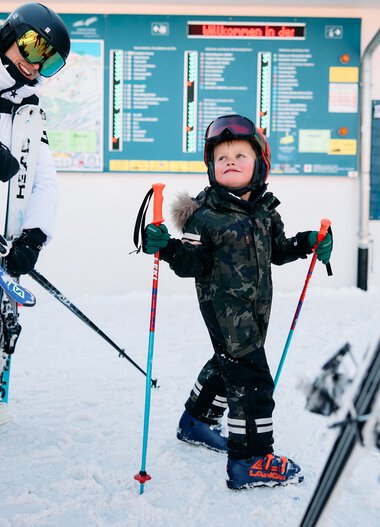 Kinder im Skigebiet mit Skischuhen und Skiern, im Hintergrund ein Informationsbildschirm am Skilift. Dachschnee und Wintersport-Atmosphäre. | © Kleinwalsertal Tourismus eGen • Fotograf: Oliver Farys