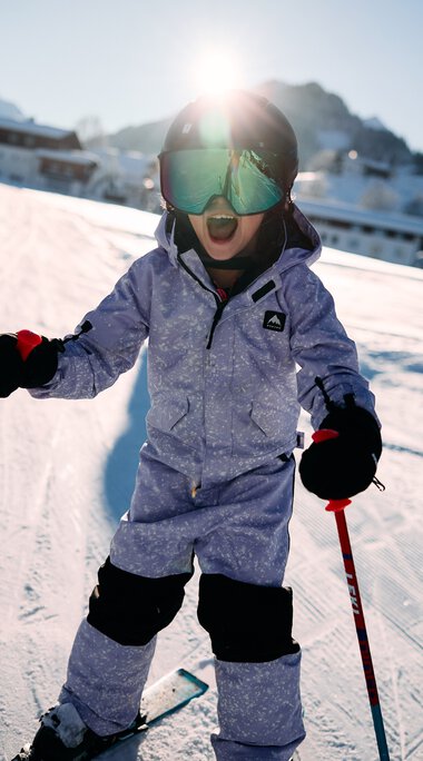 Kleines Kind beim Skifahren auf einer Piste im Snowboard- und Skigebiet im Kleinwalsertal, umgeben von Bergen, mit Skiern und -stöcken, trägt Skibrille und Winterkleidung. | © Kleinwalsertal Tourismus eGen • Fotograf: Oliver Farys