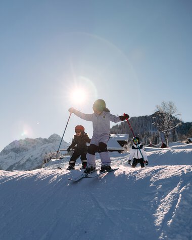 Kinder beim Skifahren auf einer schneebedeckten Piste in den Bergen bei Sonnenlicht, begleitet von einem Erwachsenen. | © Kleinwalsertal Tourismus eGen • Fotograf: Oliver Farys