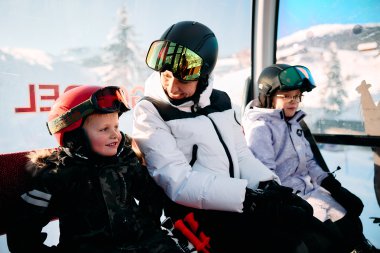 Ein Erwachsener mit zwei Kindern sitzt in einer Gondel auf dem Ski-Transport, trägt Skihelme und Skibrillen, im Hintergrund verschneite Landschaft. | © Kleinwalsertal Tourismus eGen • Fotograf: Oliver Farys