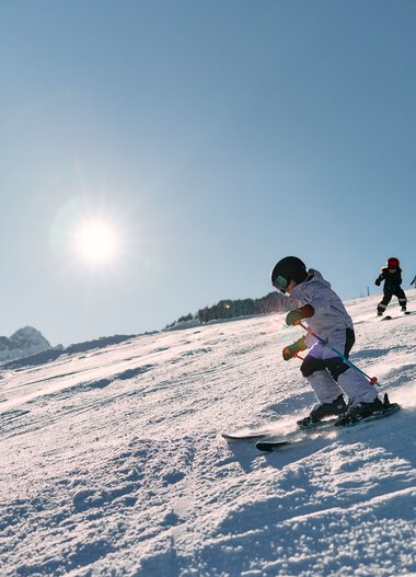 Kleines Kind beim Skifahren auf einer schneebedeckten Piste, sonniger Tag, andere Kinder im Hintergrund, Berg im Hintergrund. | © Kleinwalsertal Tourismus eGen • Fotograf: Oliver Farys