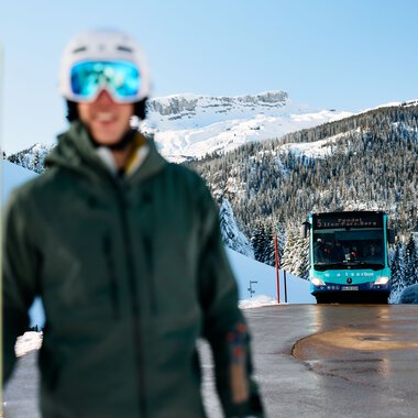Mensch mit Ski-Ausrüstung und Helm steht vor den Bergen im winterlichen Kleinwalsertal, im Hintergrund ein Ski-Bus auf der Straße. | © Kleinwalsertal Tourismus eGen • Fotograf: Oliver Farys
