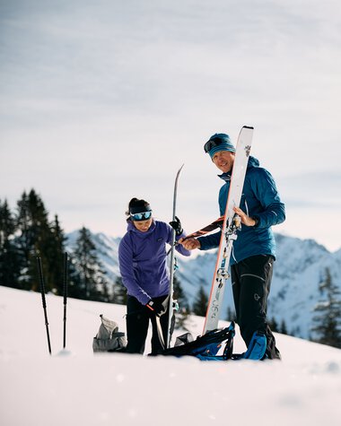 Zwei Skifahrer auf einer verschneiten Berglandschaft, einer hilft dem anderen mit Skiausrüstung, im Hintergrund Berge und Tannen. | © Kleinwalsertal Tourismus eGen • Fotograf: Oliver Farys