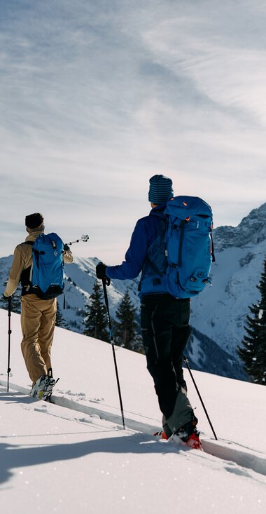 Three skitourers ascending in snowy mountain landscape in Kleinwalsertal, surrounded by mountains and pine trees, sunny winter weather. | © Kleinwalsertal Tourismus eGen • Photographer: Oliver Farys