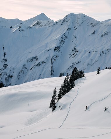 Winterlandschaft in den Bergen mit Schneespuren und Skifahrern im Kleinwalsertal, beeindruckender Ausblick auf verschneite Gipfel und Berge. | © Kleinwalsertal Tourismus eGen • Fotograf: Oliver Farys