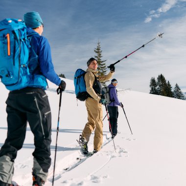 Drei Skitourengeher mit Rucksäcken in Schneelandschaft, einer macht eine selfie-artige Aufnahme während einer Wintertour im Kleinwalsertal. | © Kleinwalsertal Tourismus eGen • Fotograf: Oliver Farys